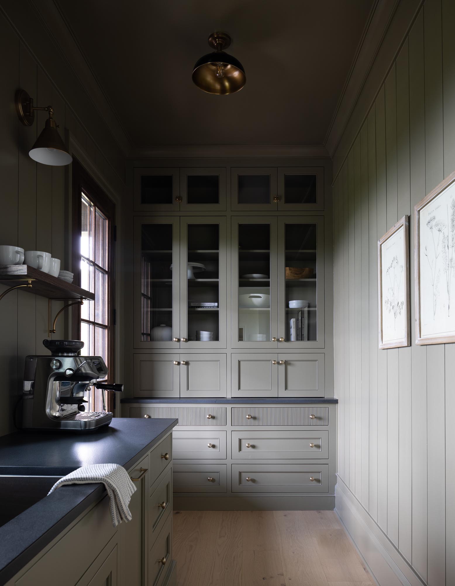 Finished scullery with Benjamin Moore Crownsville Gray cabinetry and vertical paneling, glass-front cupboards, dark counters, a coffee machine, and light wood flooring.