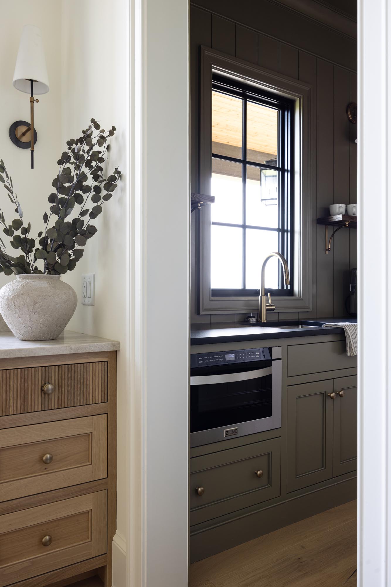 Scullery sink under a window with brass faucet and ZLINE microwave drawer, seen from the white oak kitchen